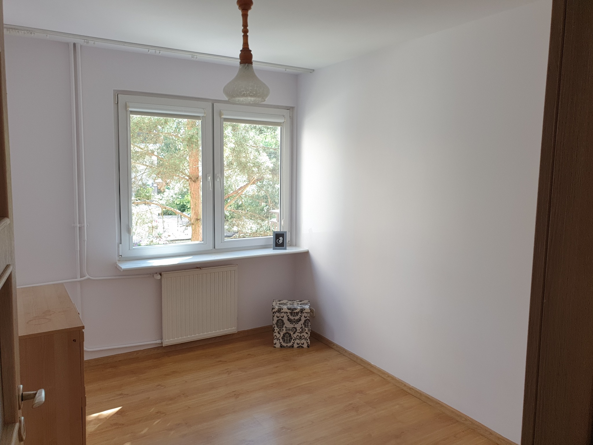 Jasne, puste wnętrze po malowaniu w Kole. Widoczne okno z widokiem na zieleń, drewniana podłoga i grzejnik. Świeżo odmalowane ściany, gotowe do aranżacji.
