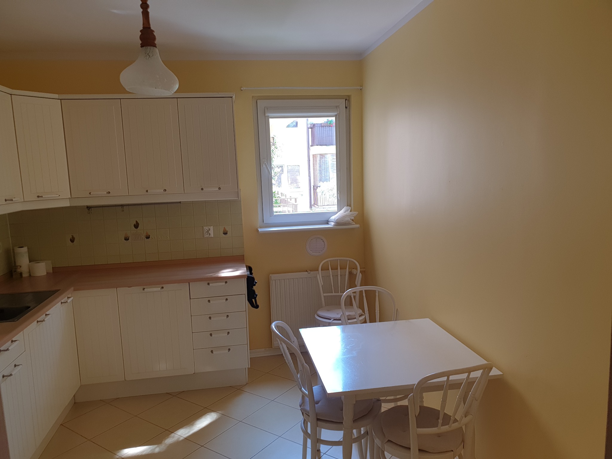 Jasna kuchnia z białymi meblami, stołem i krzesłami w stylu retro. Ściany pomalowane na żółto. Widoczne okno z widokiem na zewnątrz i lampa wisząca.