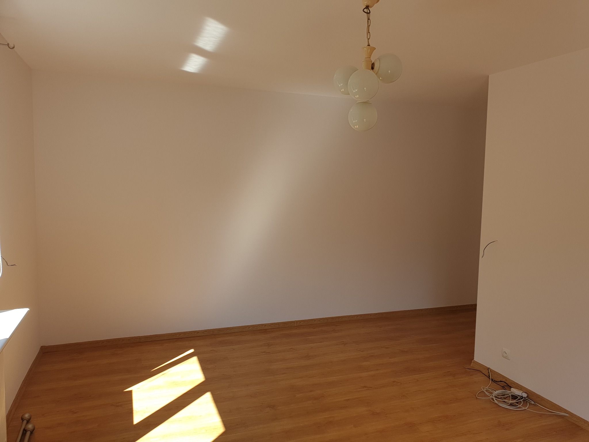 Pusty pokój po remoncie: świeżo pomalowane ściany w odcieniach bieli, parkiet, wisząca lampa w stylu retro i kable przy gniazdku. Jasne wnętrze, gotowe do umeblowania.