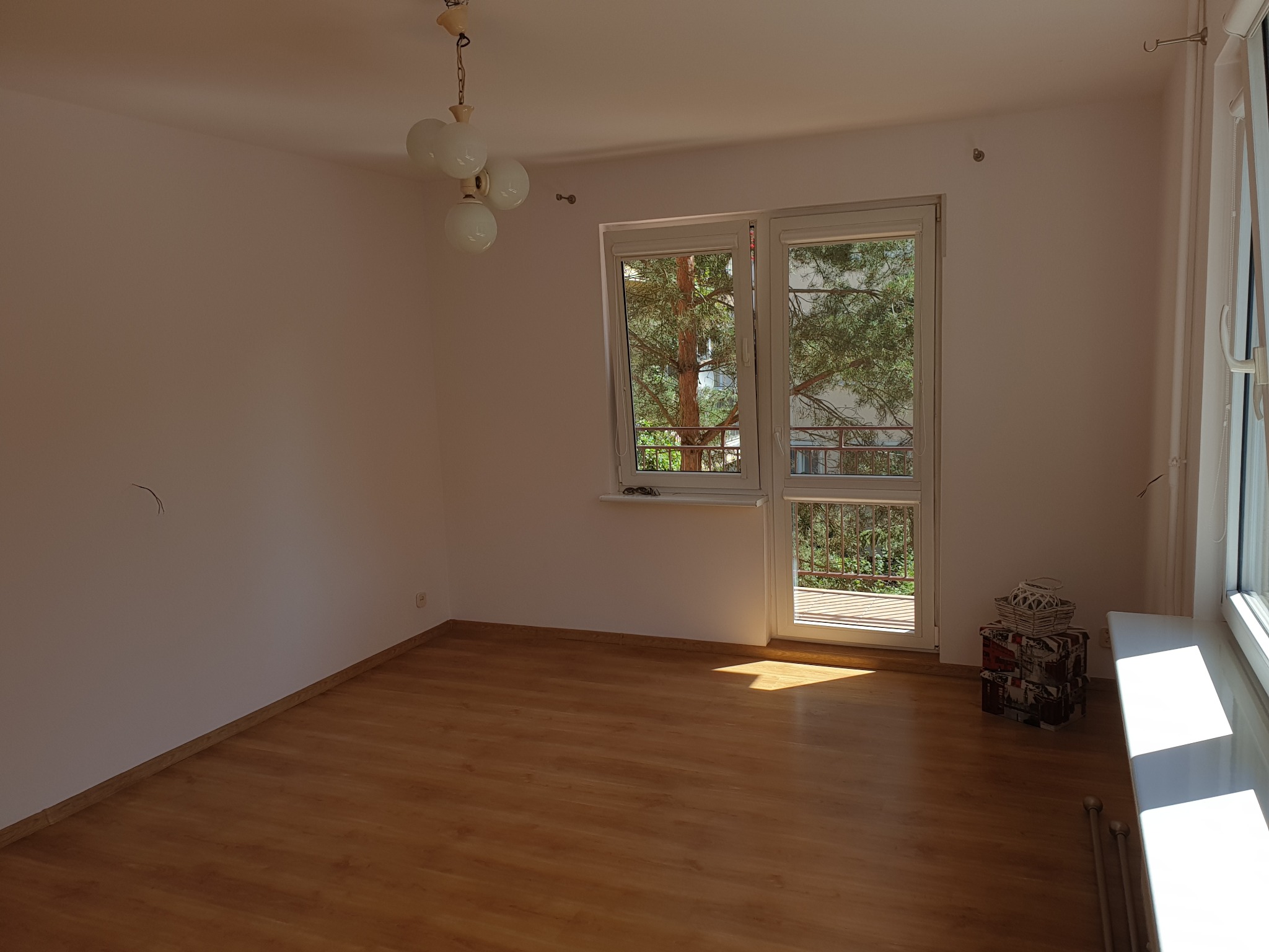 Jasne wnętrze pokoju z drewnianą podłogą, widokiem na zieleń za oknem i lampą w stylu retro. Ściany pomalowane na biało. Dekoracyjne pudełka pod oknem.