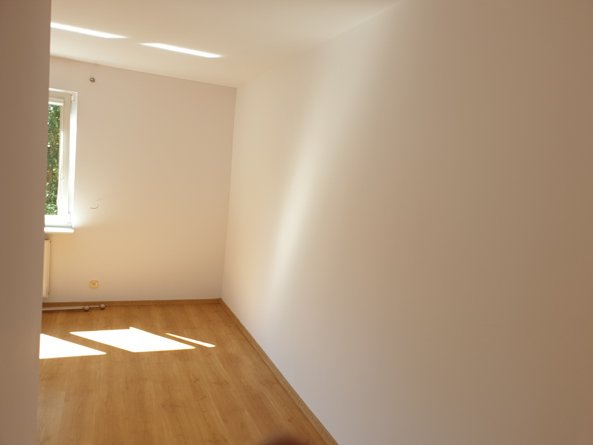Pusty, jasny pokój z parkietem i świeżo pomalowanymi ścianami w odcieniach beżu. Promienie słoneczne wpadają przez okno, tworząc geometryczne wzory na podłodze.