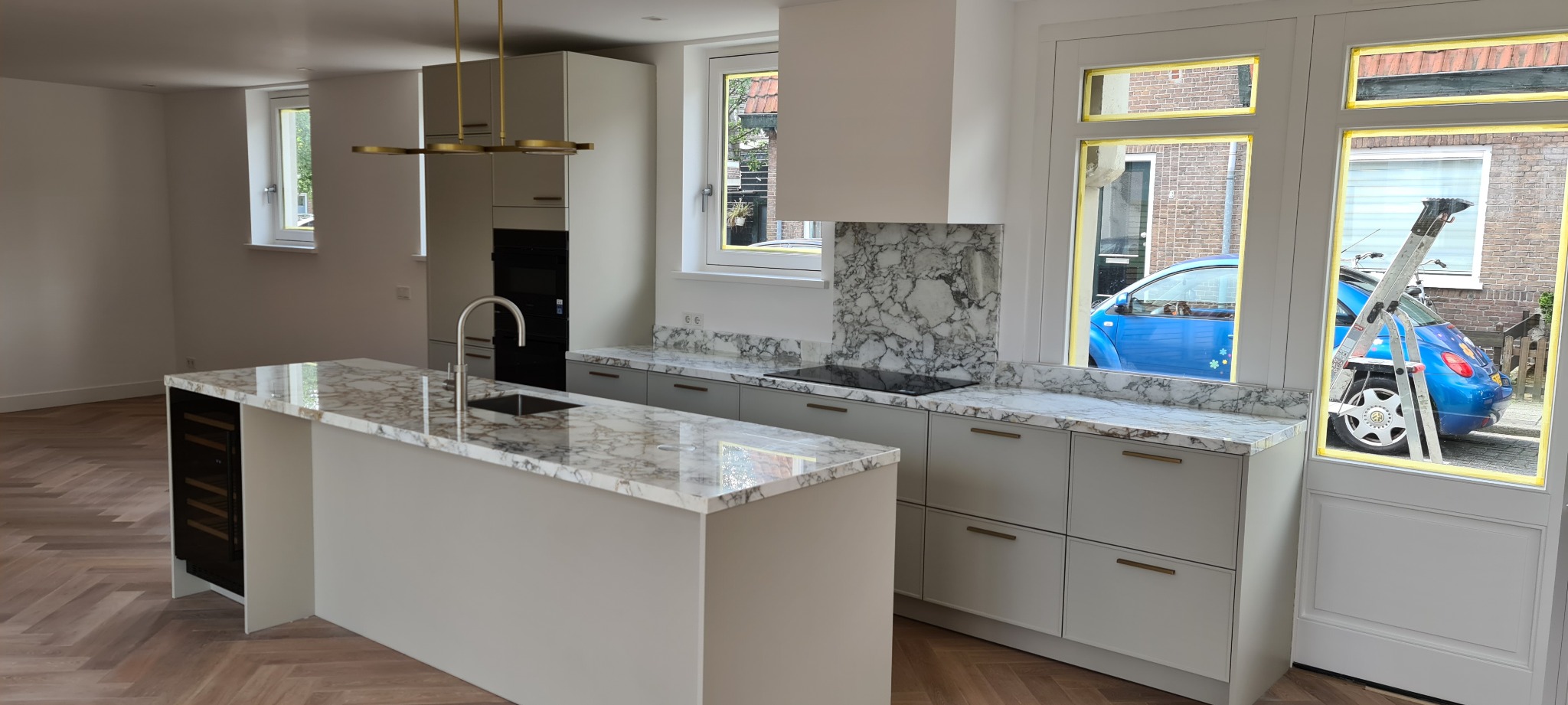 Nowoczesna kuchnia z marmurowym blatem, wyspą i parkietem w jodełkę. Okno z taśmą malarską, drabina i fragment niebieskiego auta w tle.