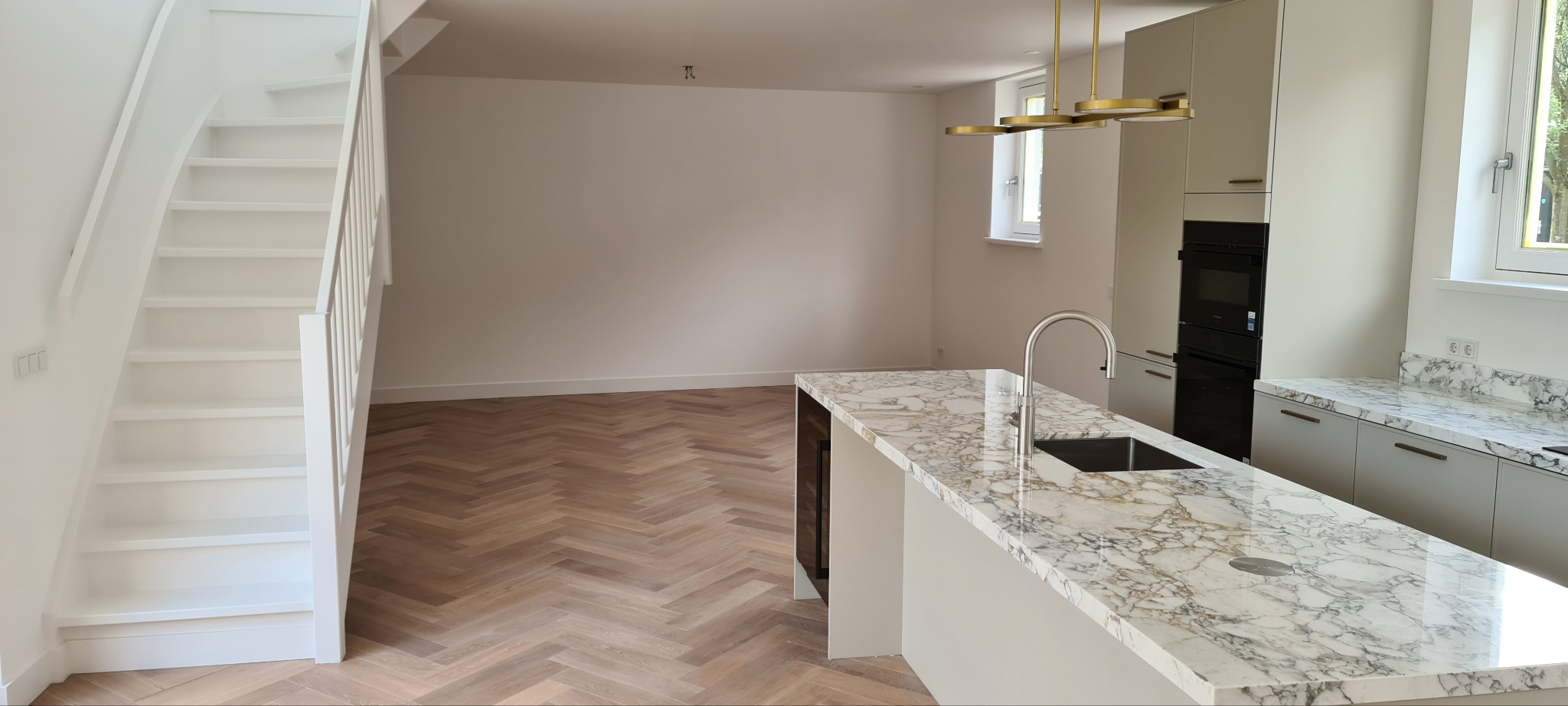 Nowoczesna kuchnia z marmurowym blatem i parkietem w jodełkę, widok na białe schody i minimalistyczne oświetlenie. Jasne wnętrze po remoncie.