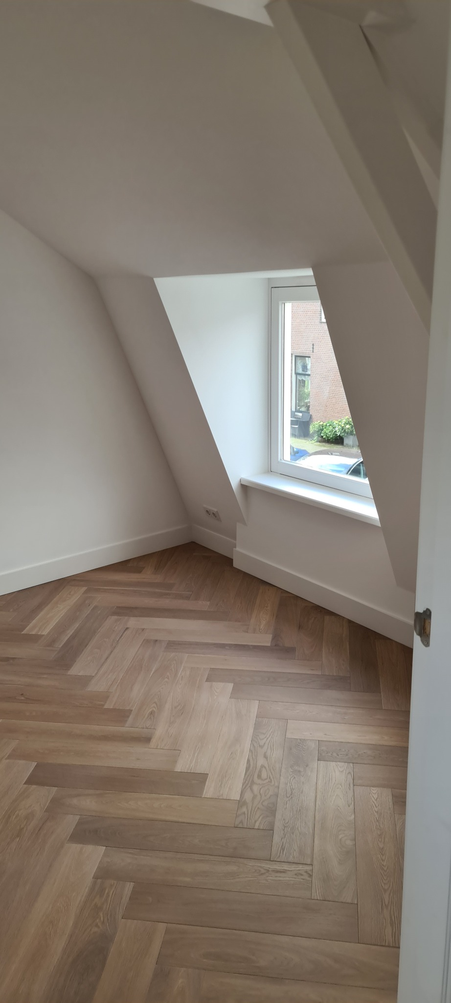 Jasne, przestronne poddasze z oknem i parkietem ułożonym w jodełkę. Ściany i sufit pomalowane na biało. Widoczny fragment drzwi po prawej stronie.