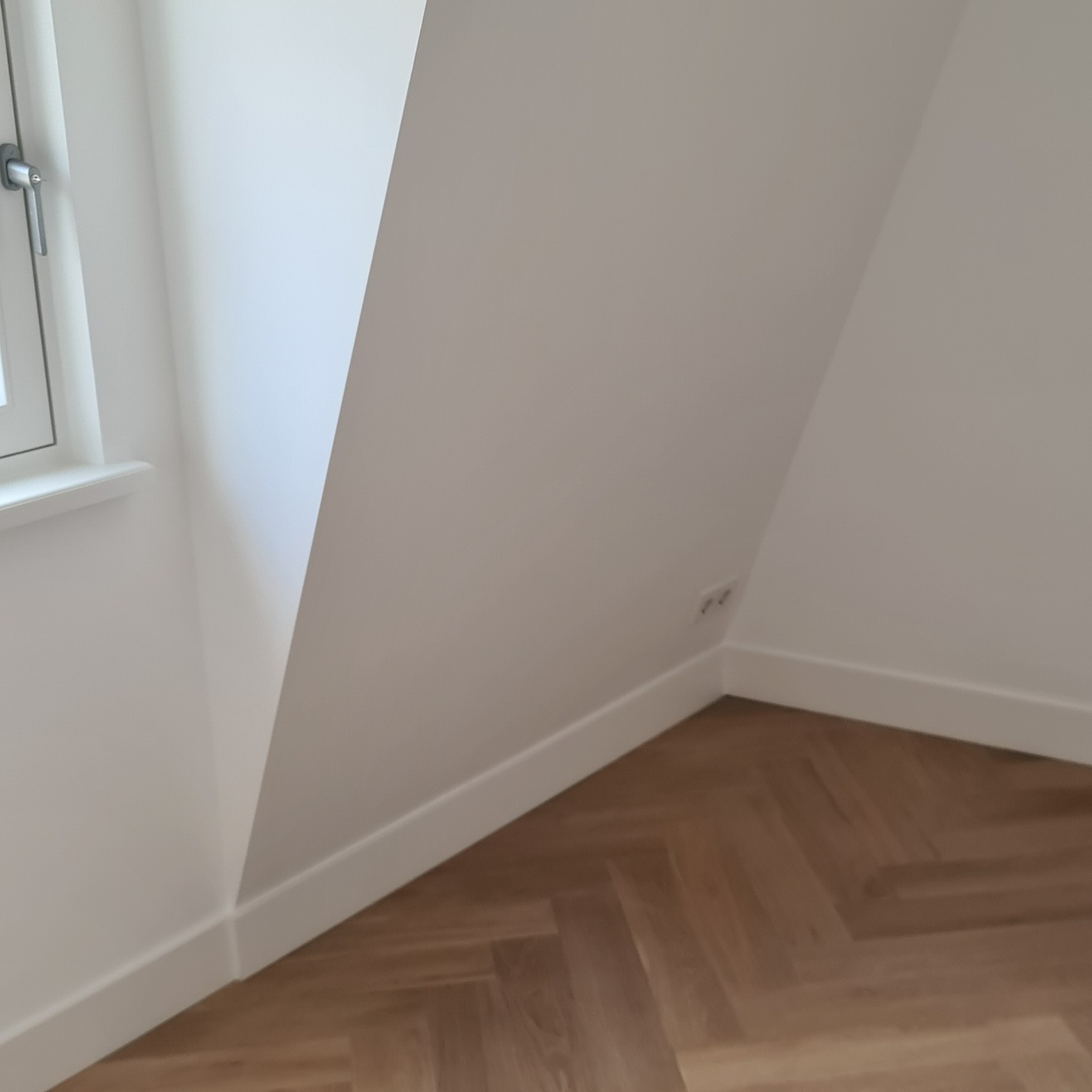 Jasne wnętrze poddasza po wykończeniu: parkiet w jodełkę, białe ściany i listwy przypodłogowe, okno z widocznym fragmentem ramy. Gniazdka elektryczne przy podłodze.