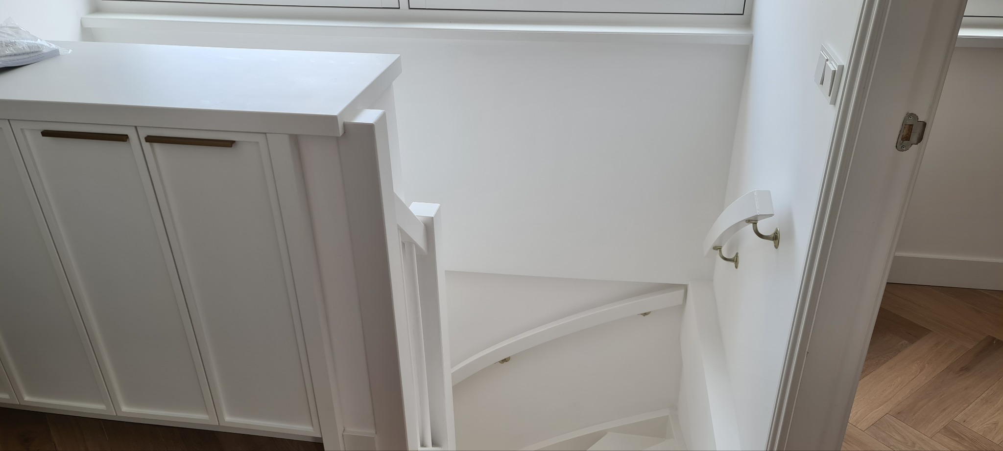 Białe, drewniane schody kręte z poręczą i szafką z złotymi uchwytami, w jasnym wnętrzu. Widok z góry na dół.