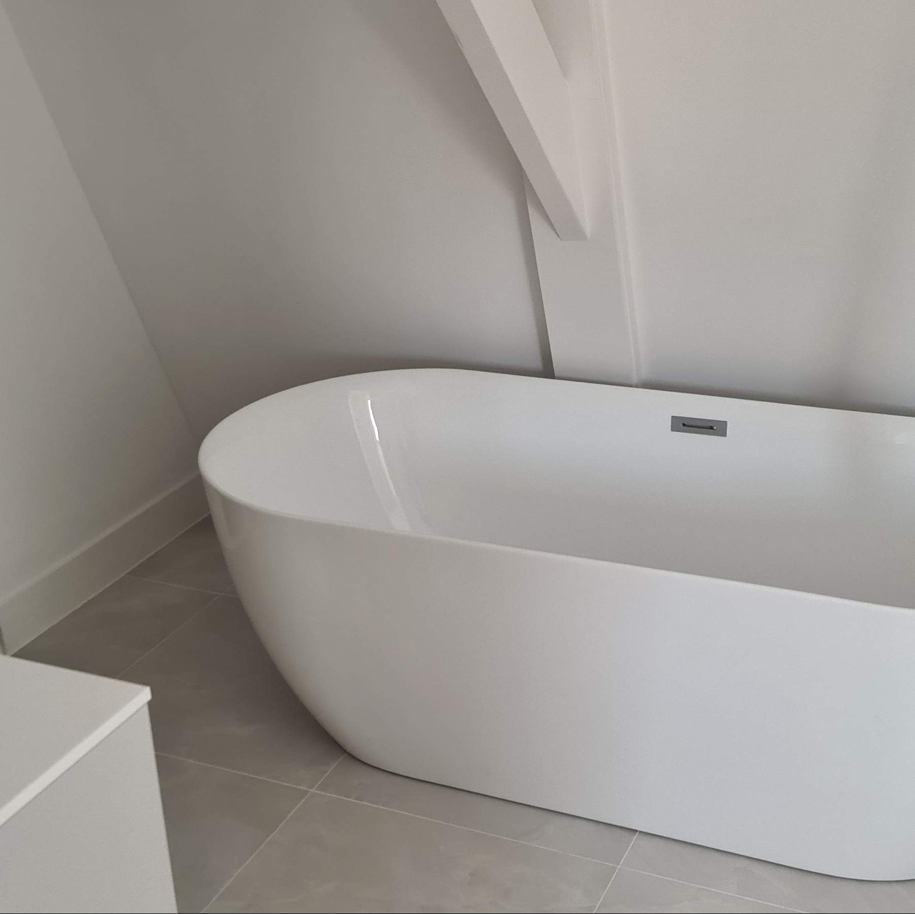 Nowoczesna, wolnostojąca wanna w minimalistycznej łazience z szarą podłogą i białymi ścianami. Uwagę zwraca ukośny element architektoniczny na suficie.