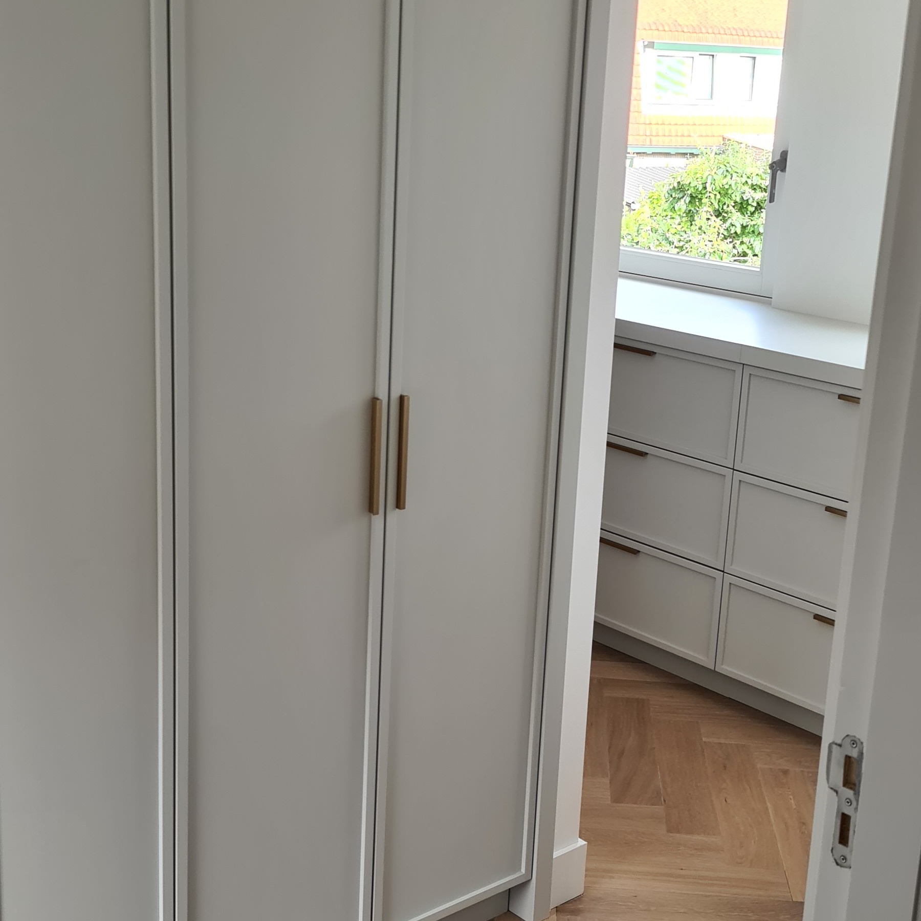 Biała szafa wnękowa z złotymi uchwytami, widok z boku na białe szafki pod oknem i parkiet ułożony w jodełkę. Subtelne detale wykończenia.