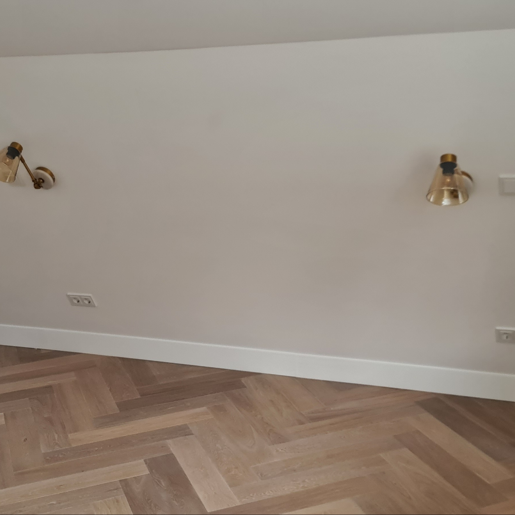 Jasny parkiet ułożony w jodełkę, białe ściany z listwami przypodłogowymi i dwa złote kinkiety. Widoczne gniazdka elektryczne. Minimalistyczne wnętrze.