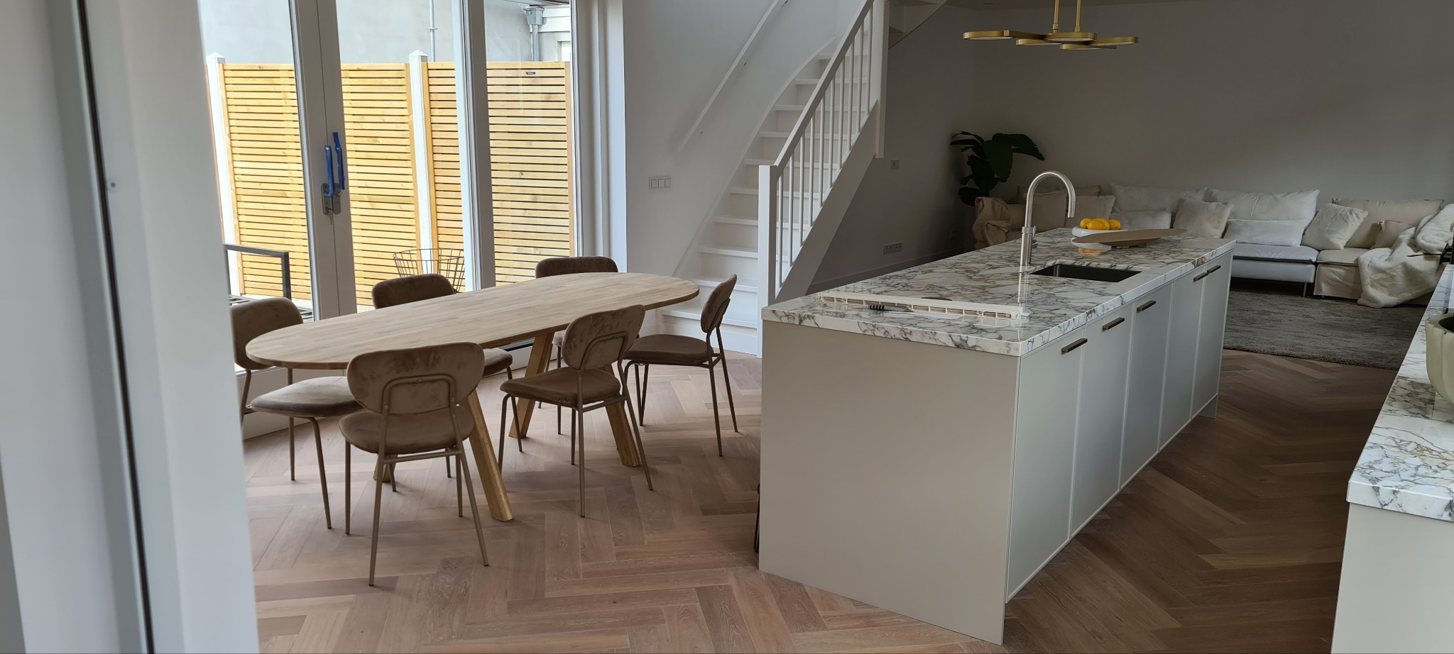 Jasne wnętrze: parkiet w jodełkę, stół z krzesłami, wyspa kuchenna z marmurowym blatem. Widoczne schody i sofa. Minimalistyczny design, naturalne światło.