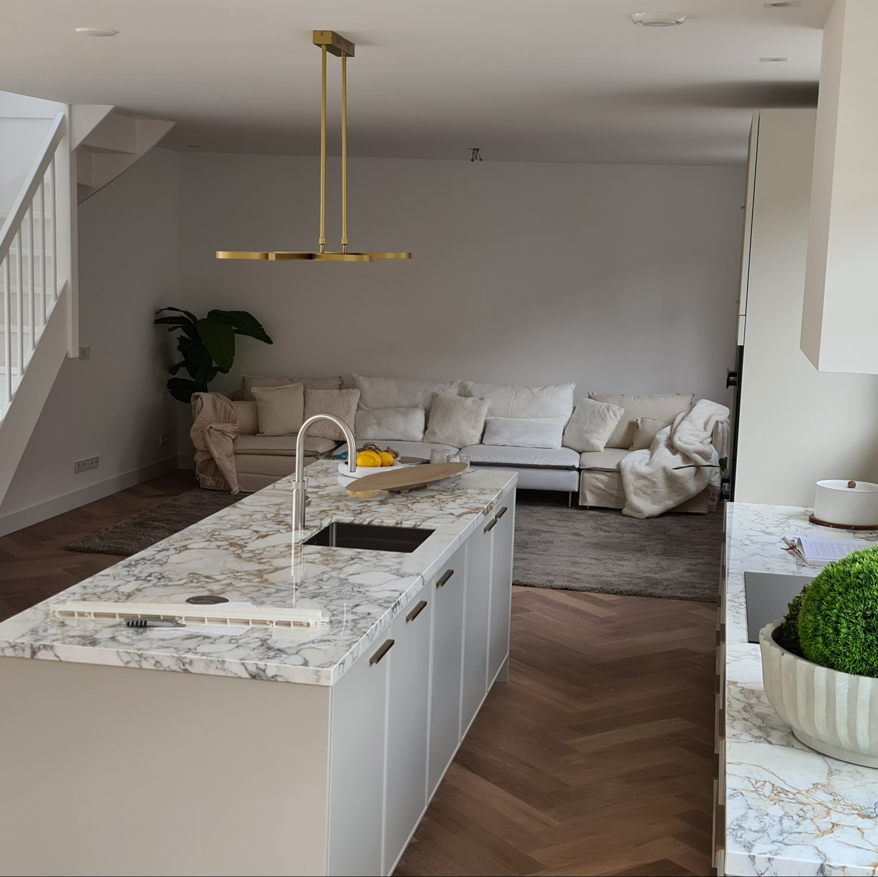 Nowoczesna wyspa kuchenna z marmurowym blatem i złotymi akcentami oświetlenia, w tle salon z sofą i schody. Parkiet ułożony we wzór jodełki.