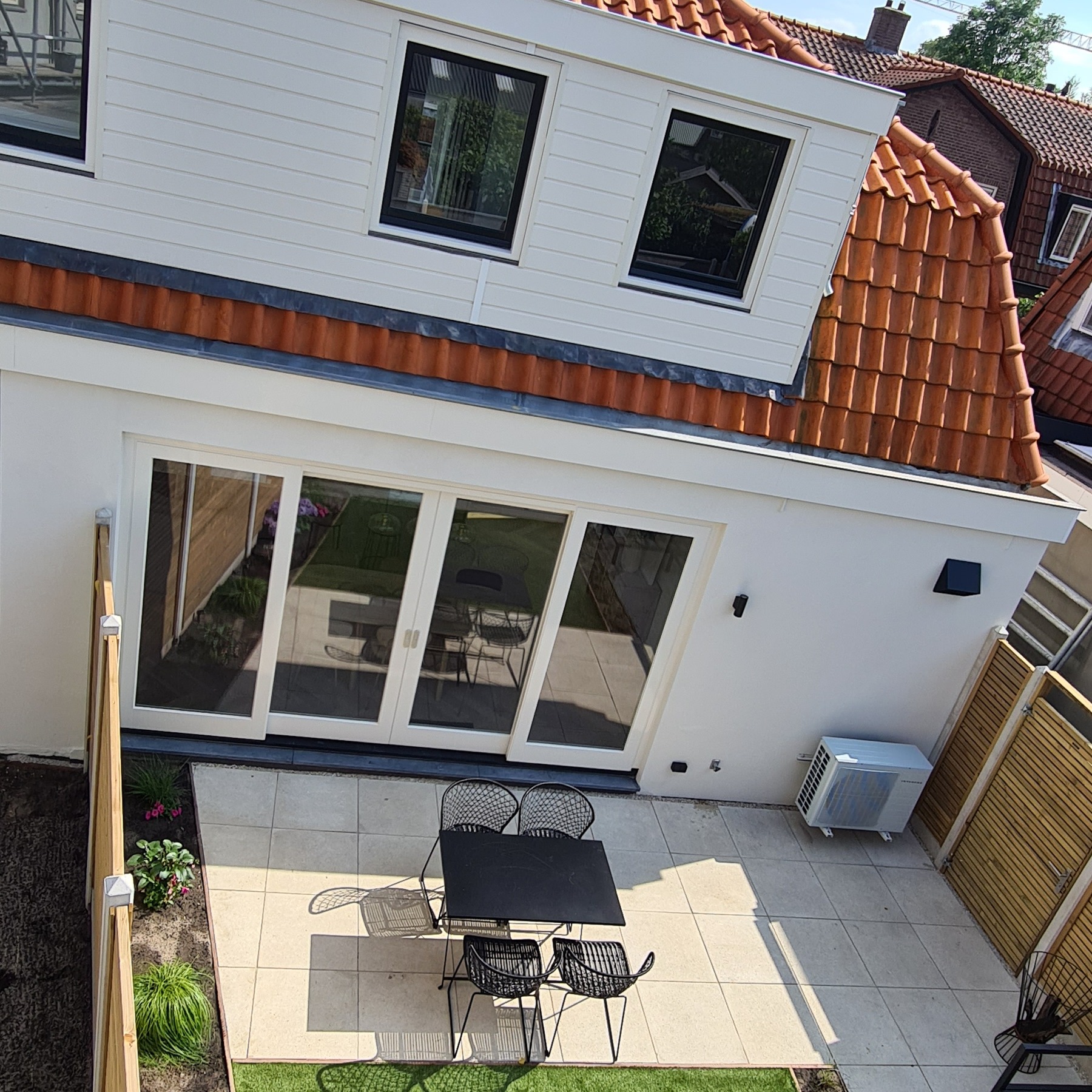 Widok z góry na odnowioną elewację domu z nowymi oknami, tarasem i meblami ogrodowymi w słoneczny dzień. Widoczny klimatyzator i fragment ogrodu.