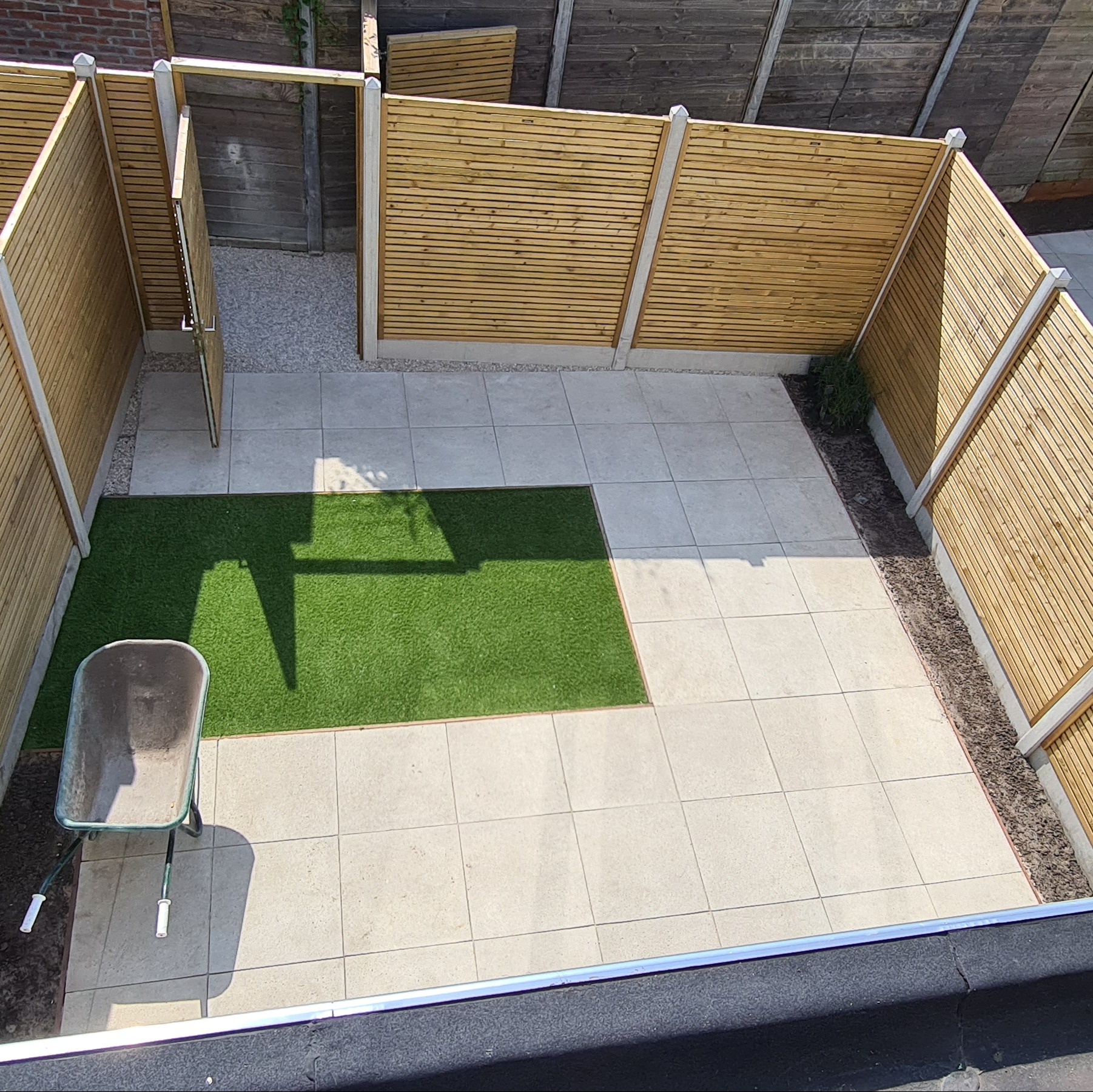 Nowoczesny taras z jasnych płyt, fragmentem sztucznej trawy i drewnianym ogrodzeniem z poziomych listew. W rogu taczka, widok z góry.
