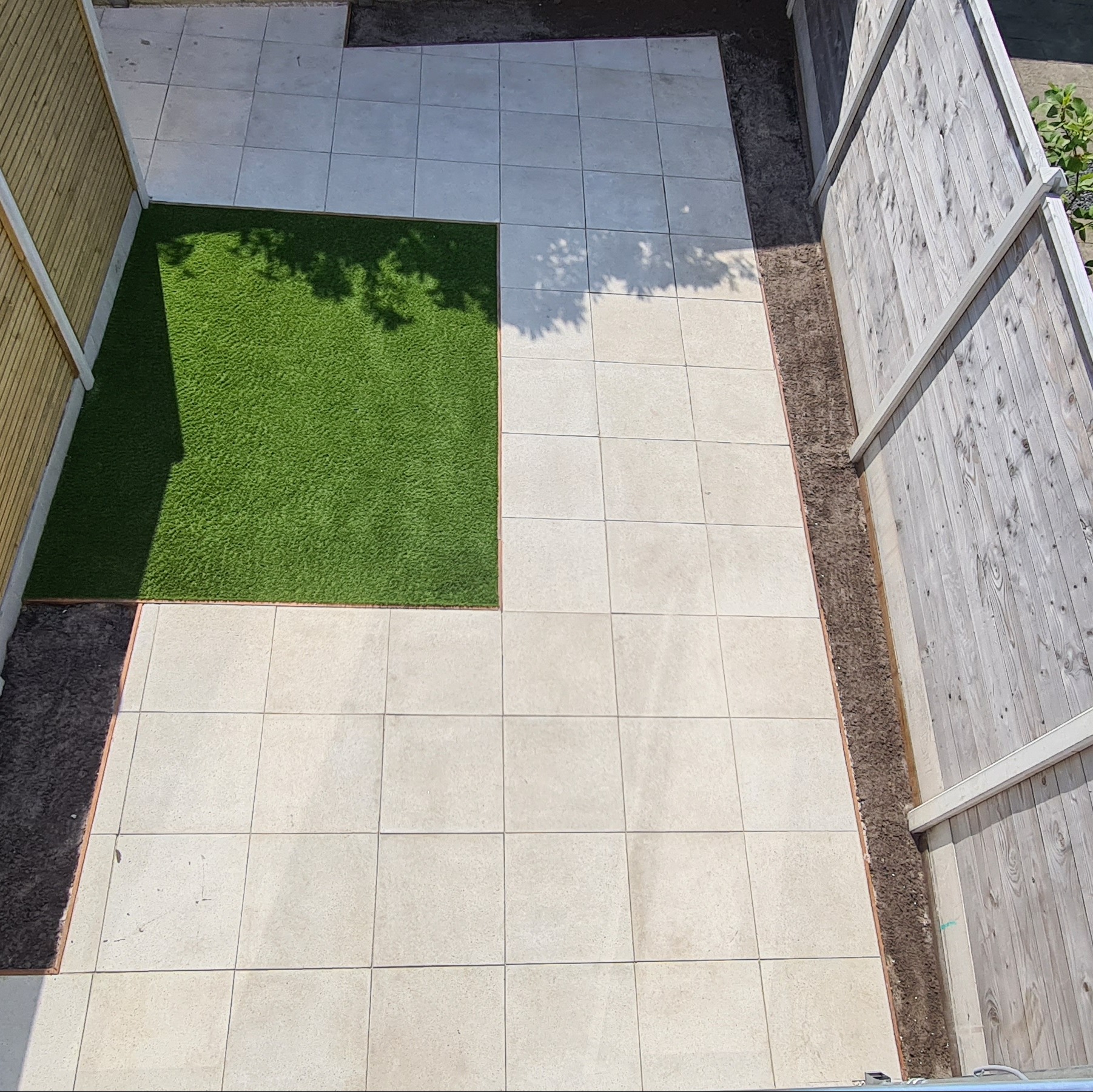 Ułożone jasne płytki tarasowe z kwadratową wstawką sztucznej trawy, widok z góry. Płot drewniany po prawej, drewniana ściana po lewej. Cień drzewa na trawie.