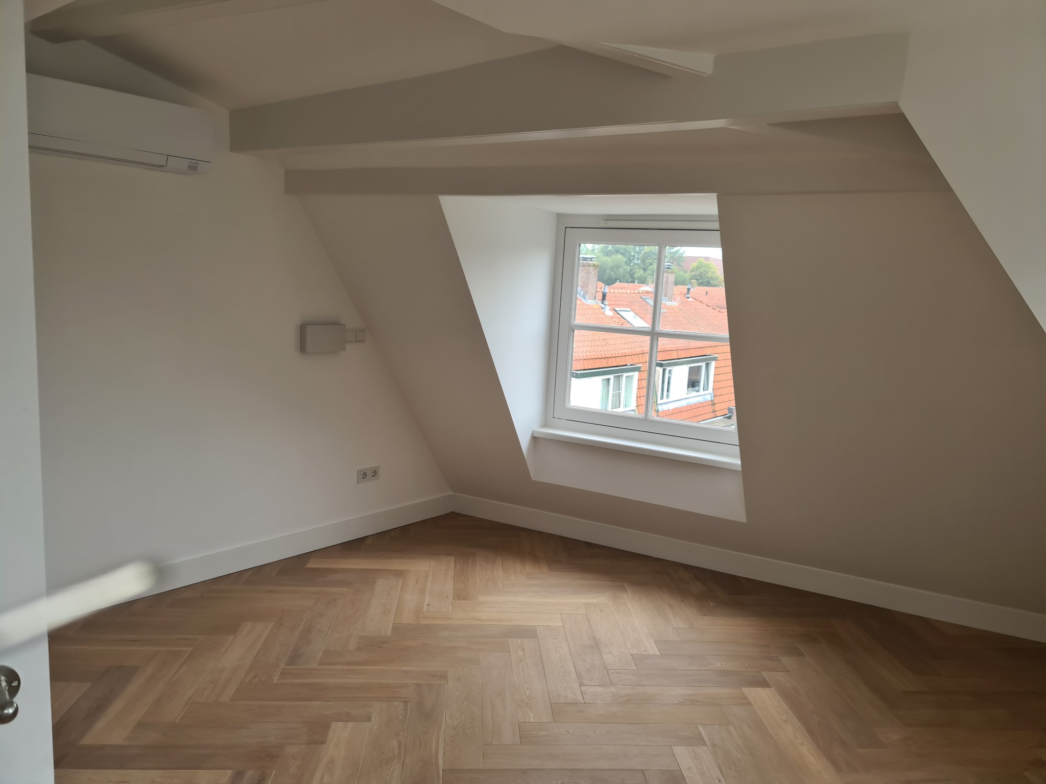 Jasne poddasze po remoncie: białe ściany, drewniana podłoga w jodełkę, okno z widokiem na dachy. Minimalistyczny design, świeżo po malowaniu.