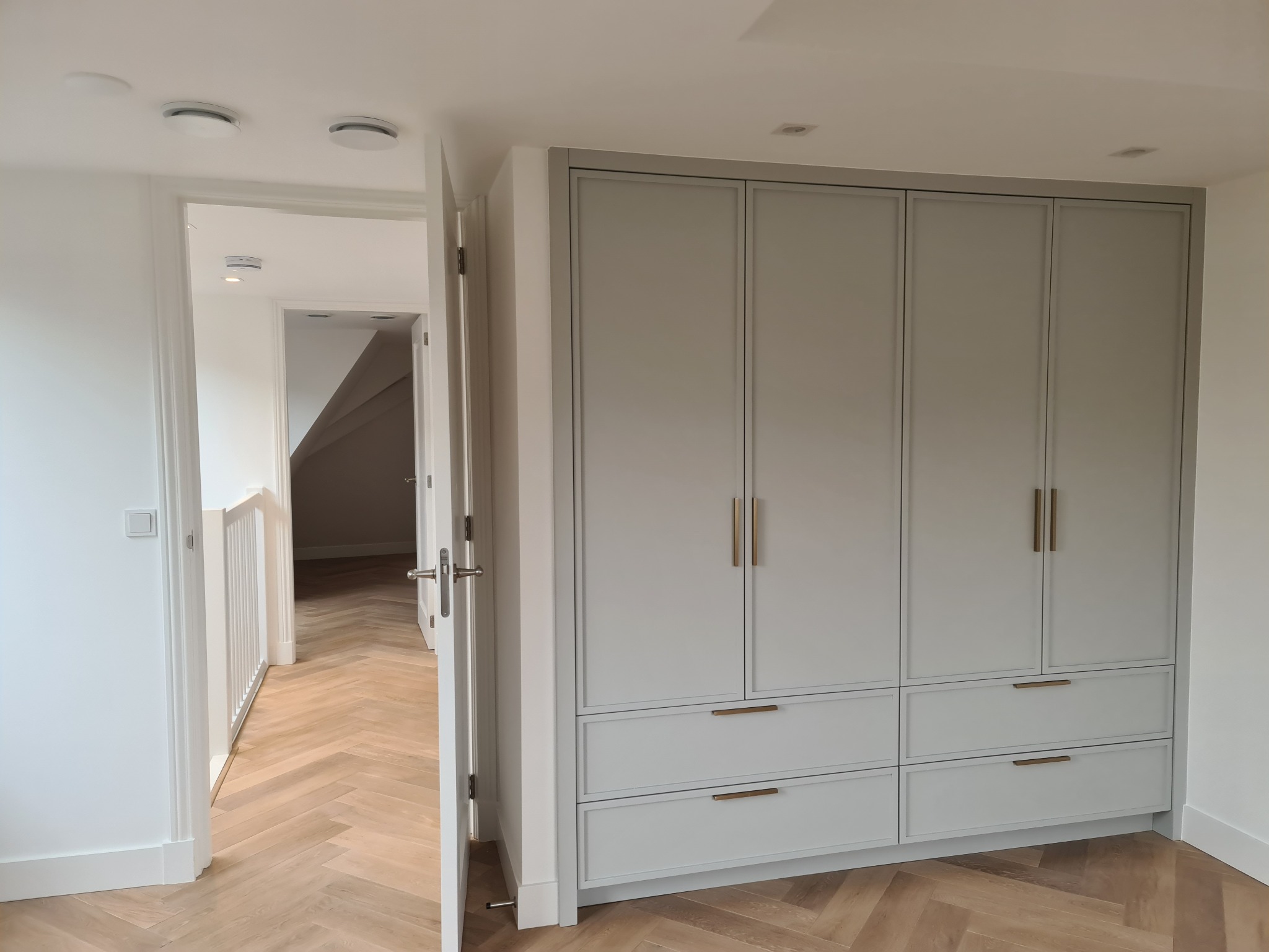 Jasnoszara szafa wnękowa z złotymi uchwytami, widok z korytarza z parkietem ułożonym w jodełkę, białe ściany i otwarte drzwi.