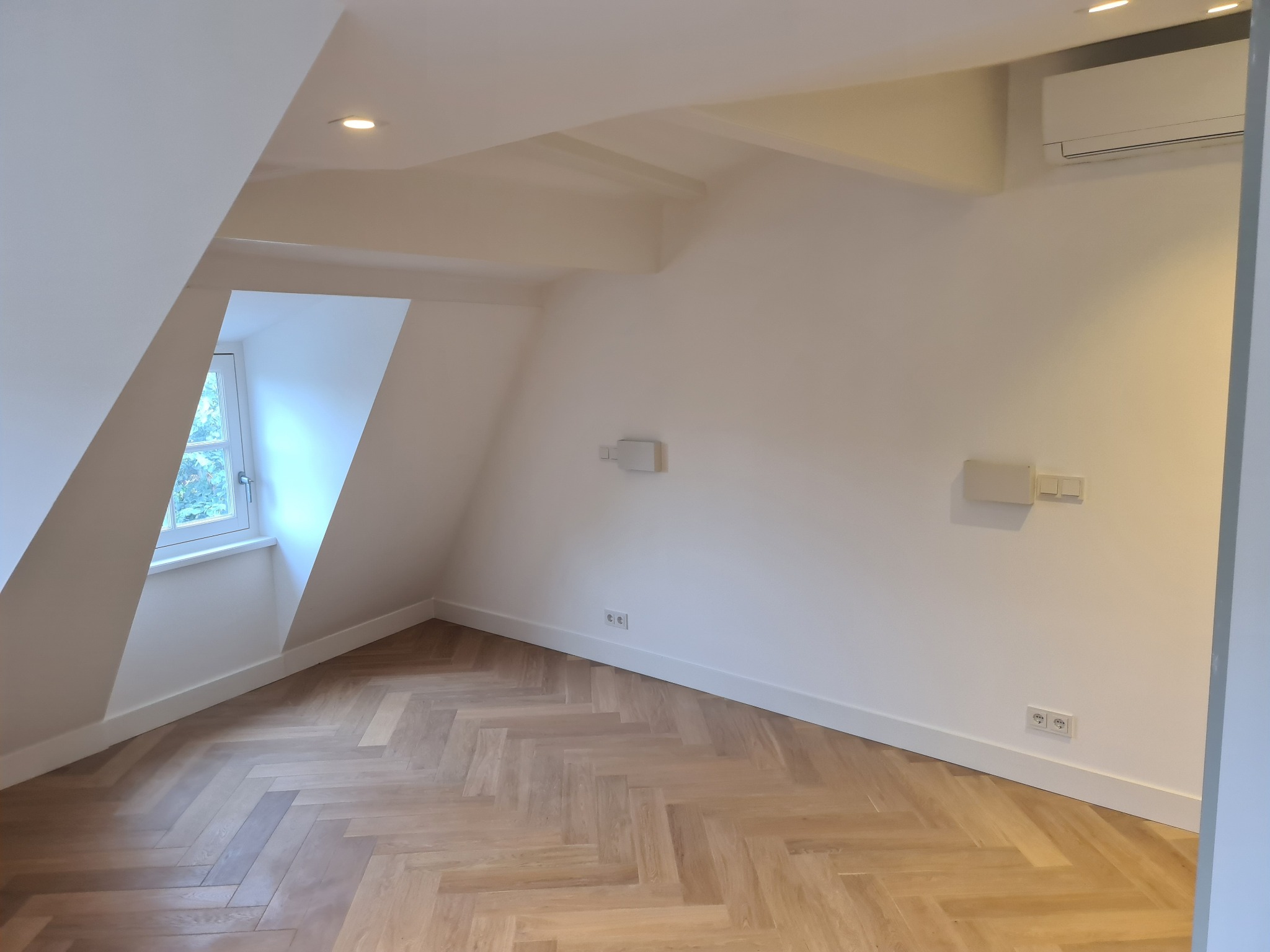 Jasne, wykończone poddasze z parkietem w jodełkę, białymi ścianami, oknem w szczycie i klimatyzacją. Widoczne gniazdka elektryczne i listwy przypodłogowe.