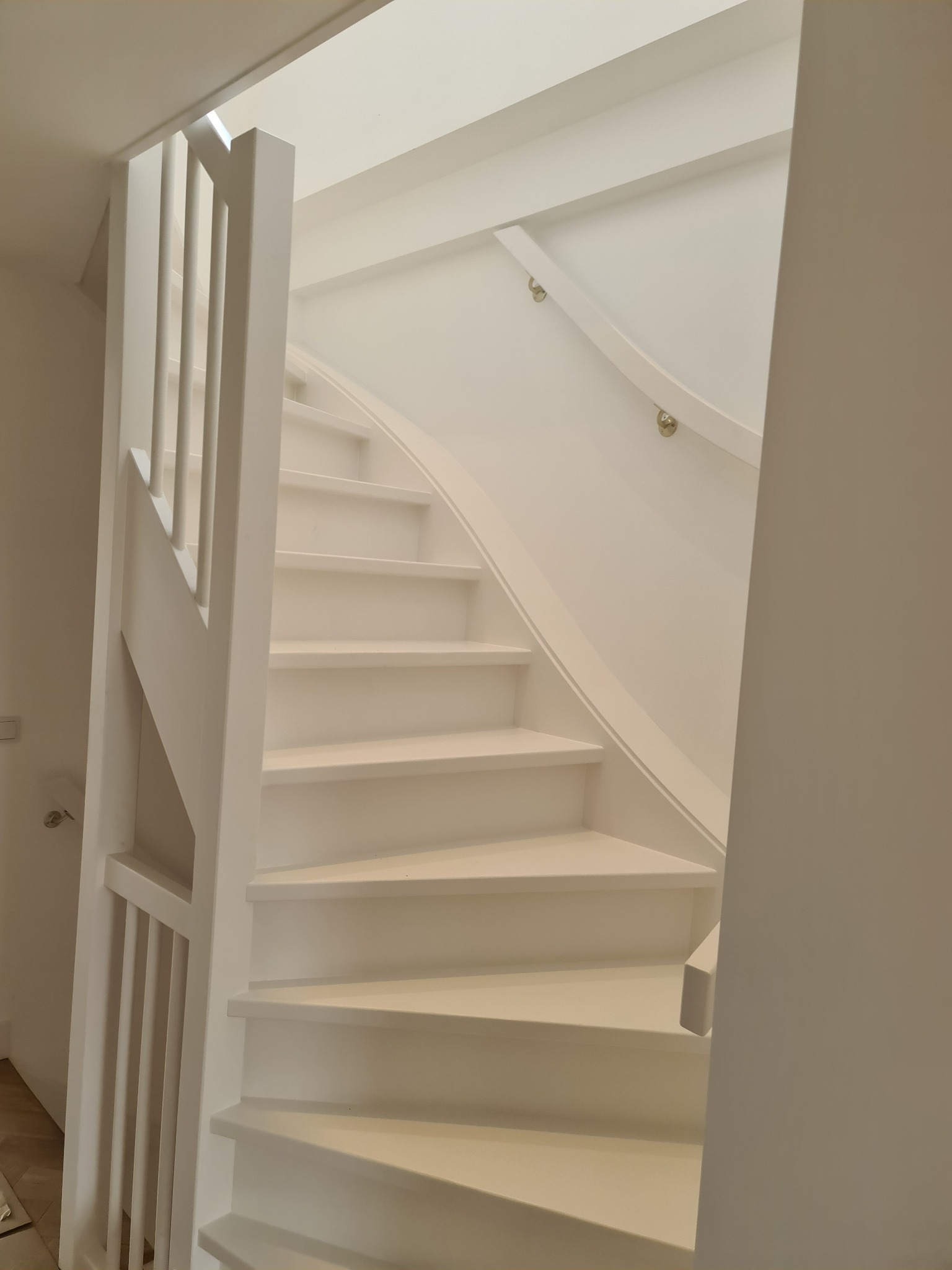 Białe, drewniane schody z poręczą i balustradą w jasnym wnętrzu. Widoczne detale mocowań poręczy. Minimalistyczny design, subtelne oświetlenie.