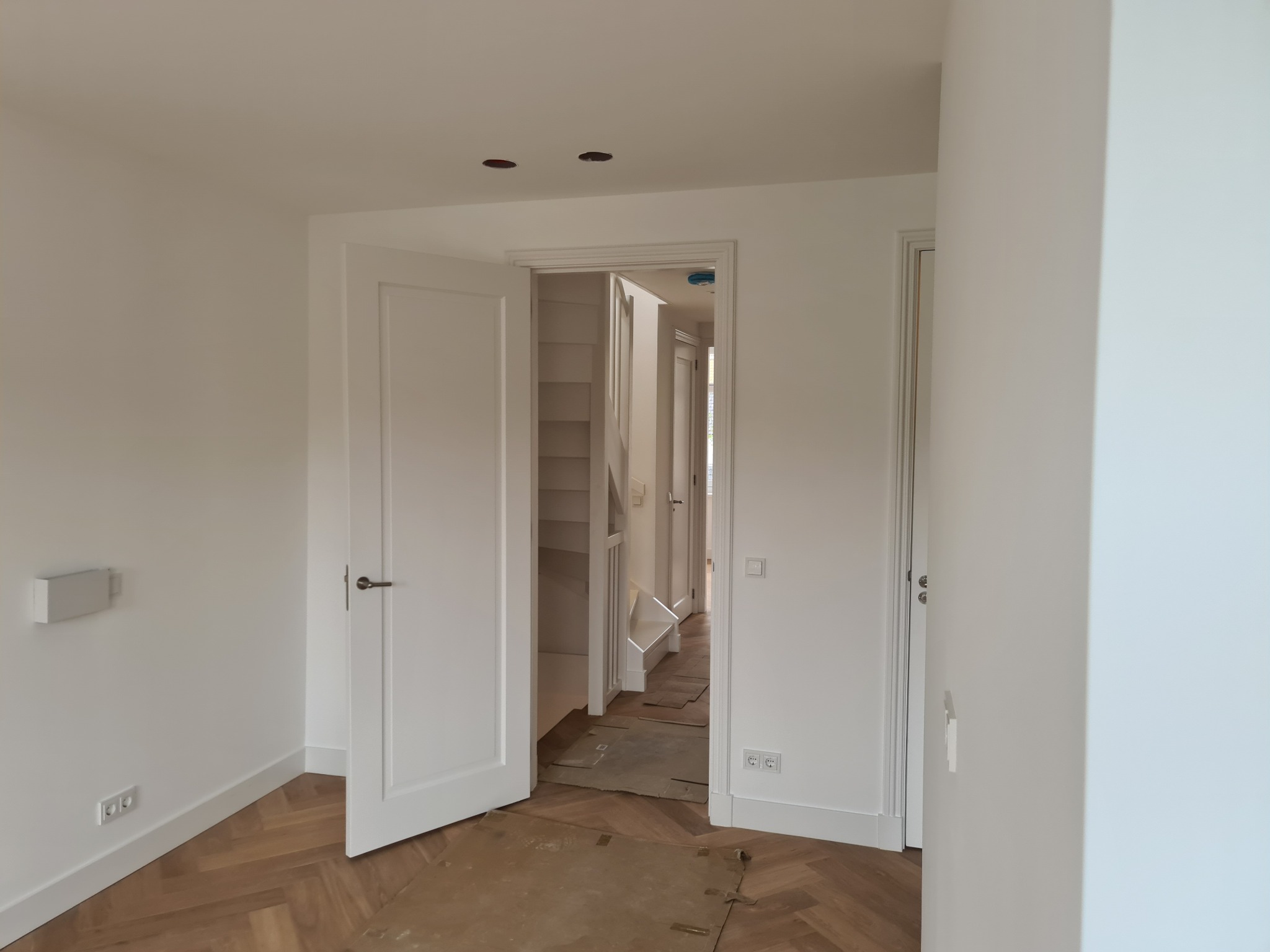 Jasne wnętrze po remoncie: białe ściany, drewniany parkiet w jodełkę zabezpieczony kartonem, otwarte białe drzwi i widok na schody. Dwa otwory w suficie.