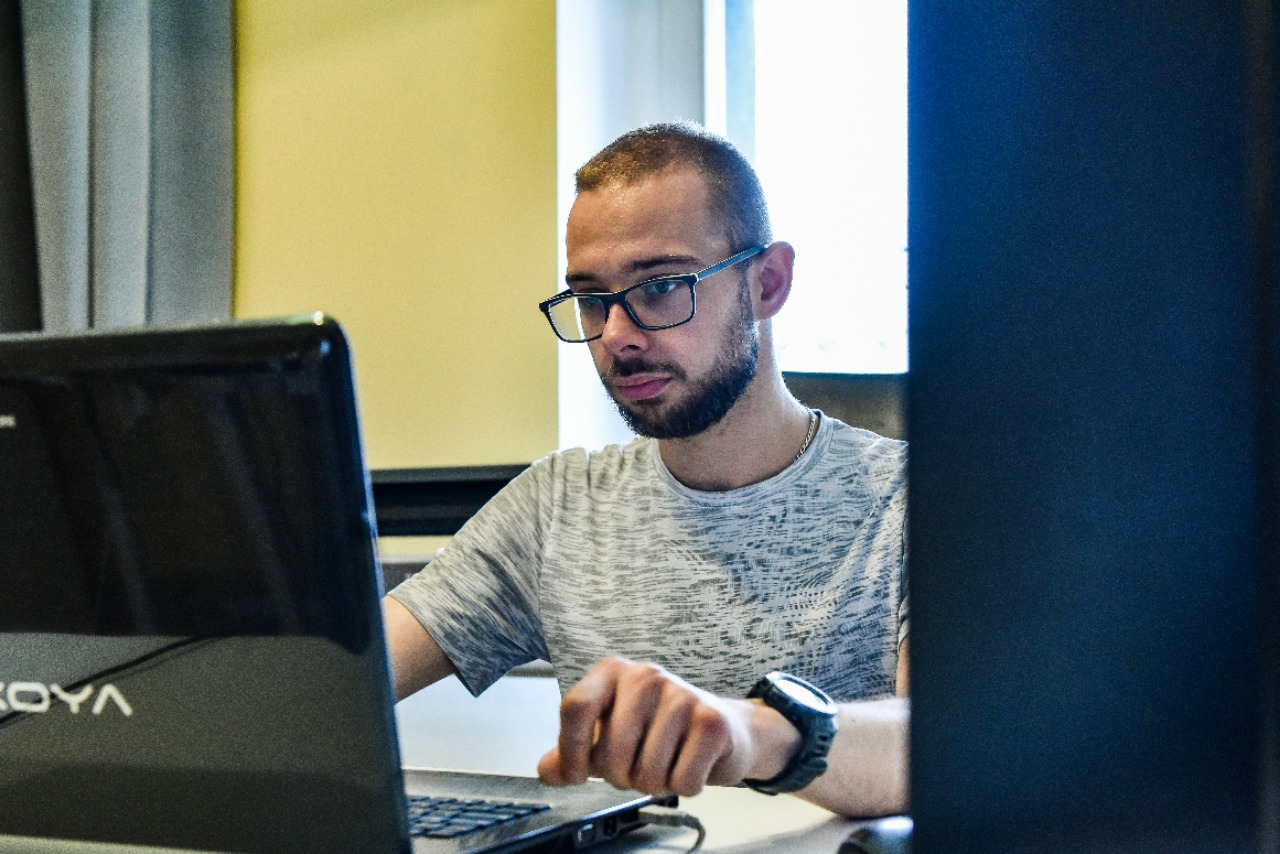 Projektant odzieży przy laptopie, skupiony na pracy, w tle żółta ściana i okno z jasnym światłem dziennym. Widoczny zegarek na ręce.