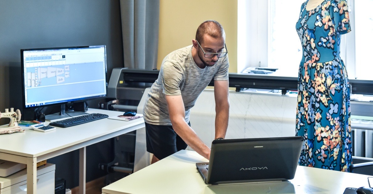 Projektant odzieży pracuje przy laptopie, obok monitor z szablonami i gotowa sukienka na manekinie, w tle ploter tnący. Jasne wnętrze pracowni.