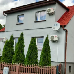 Szary dom jednorodzinny z czerwonym dachem. Na ścianach zamontowane klimatyzatory TCL i antena satelitarna nc+. W tle widoczne panele fotowoltaiczne.