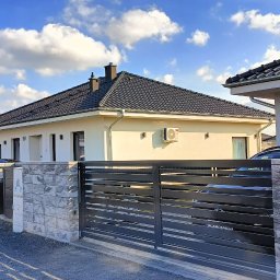 Ariel Paszczak Klimatyzacja - Nowoczesny dom jednorodzinny z klimatyzacją na elewacji, widoczny zza grafitowego ogrodzenia. Błękitne niebo z obłokami w tle. Słoneczny dzień.