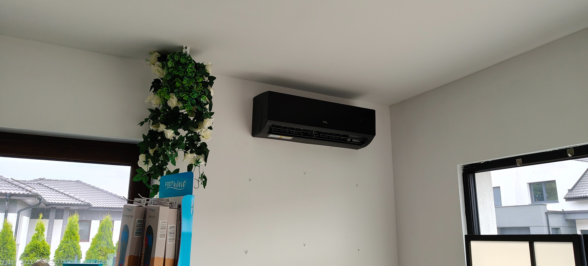 Czarna klimatyzacja TCL zamontowana na białej ścianie w nowoczesnym wnętrzu, obok okna z dekoracją kwiatową. Widoczne ślady po poprzednim montażu.