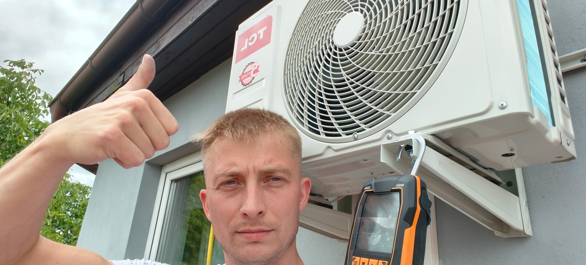 Instalator klimatyzacji TCL daje kciuk w górę obok zamontowanego urządzenia na elewacji, trzymając miernik elektroniczny. Widok z dołu na klimatyzator.
