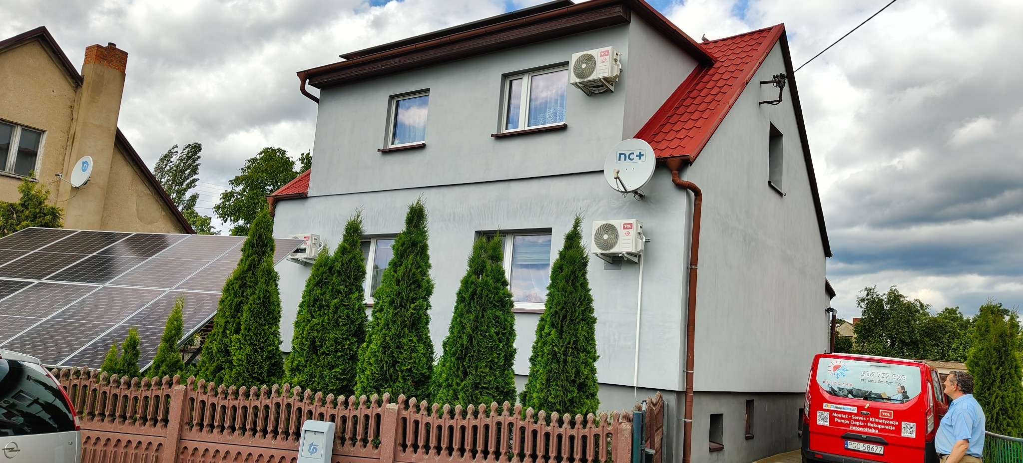Szary dom jednorodzinny z czerwonym dachem. Na ścianach zamontowane klimatyzatory TCL i antena satelitarna nc+. W tle widoczne panele fotowoltaiczne.