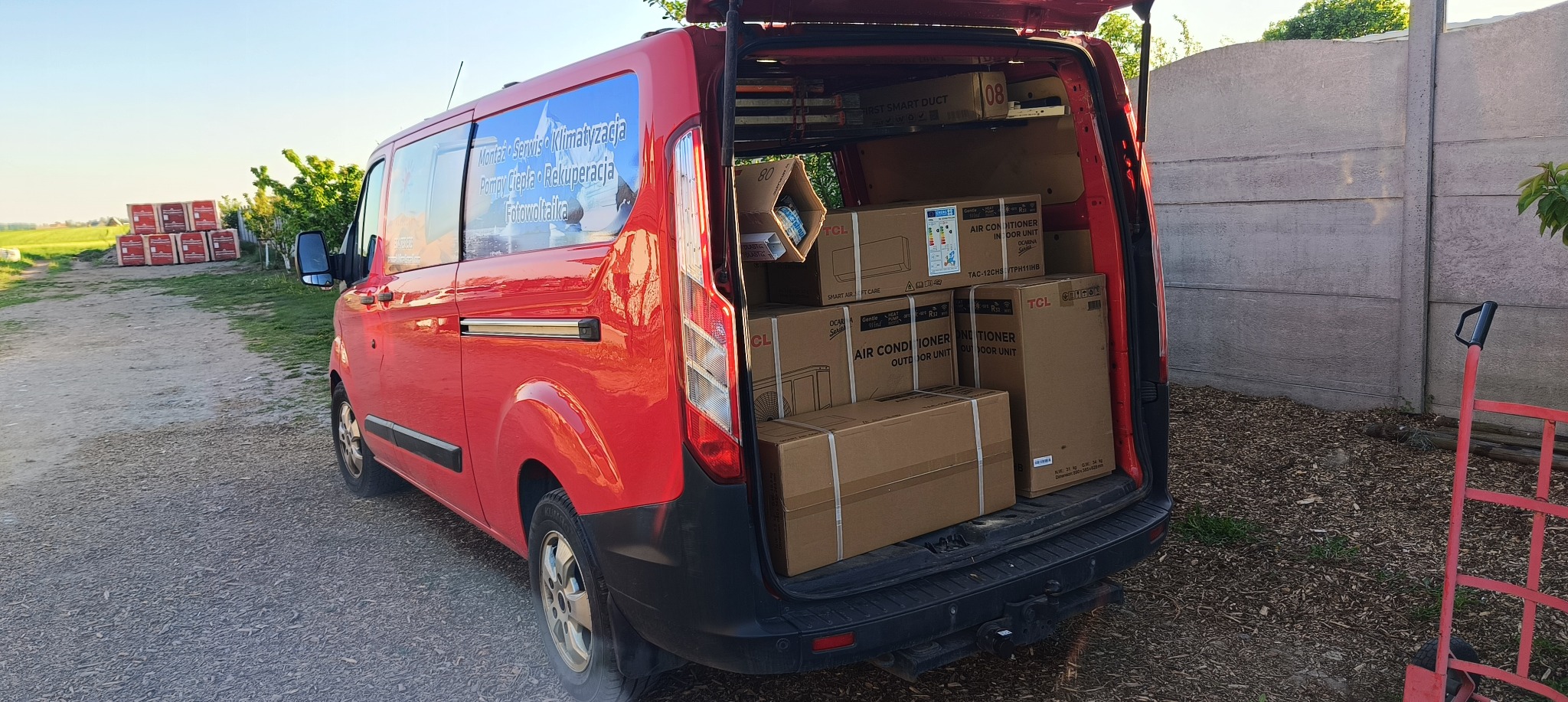 Czerwony van z otwartymi drzwiami załadowany klimatyzatorami TCL w kartonach, gotowy do montażu w Granowie. Reklama firmy na boku pojazdu.