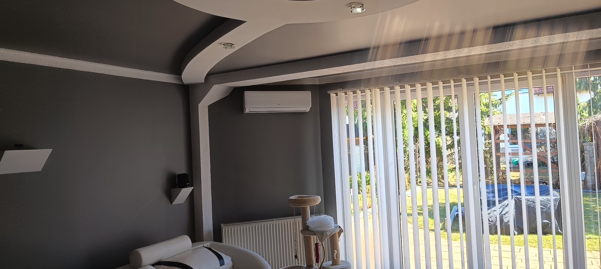 Nowoczesne wnętrze z klimatyzatorem TCL na szarej ścianie, widoczne przez okno pionowe żaluzje i zielony ogród. Dekoracyjny sufit z oświetleniem punktowym.