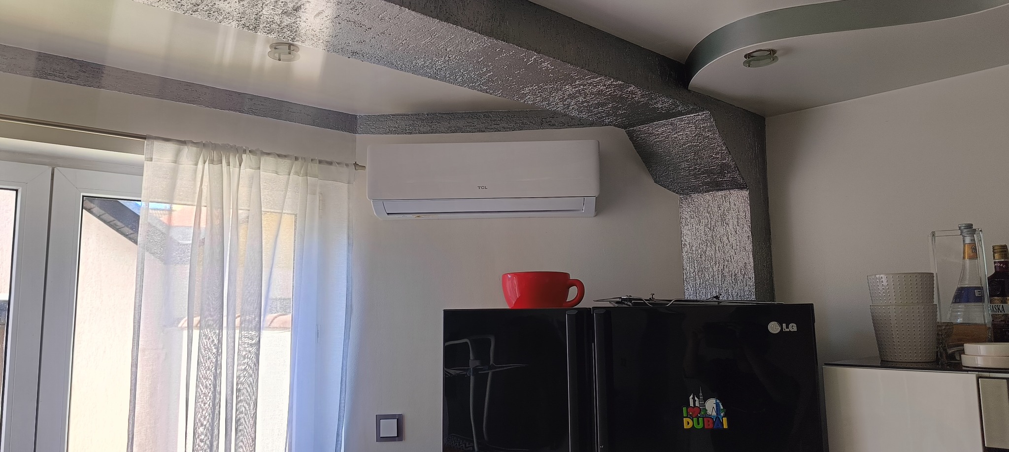 Biała klimatyzacja TCL zamontowana na ścianie w pokoju z dekoracyjnymi belkami sufitowymi i czarną lodówką LG. Czerwony kubek stoi na lodówce.