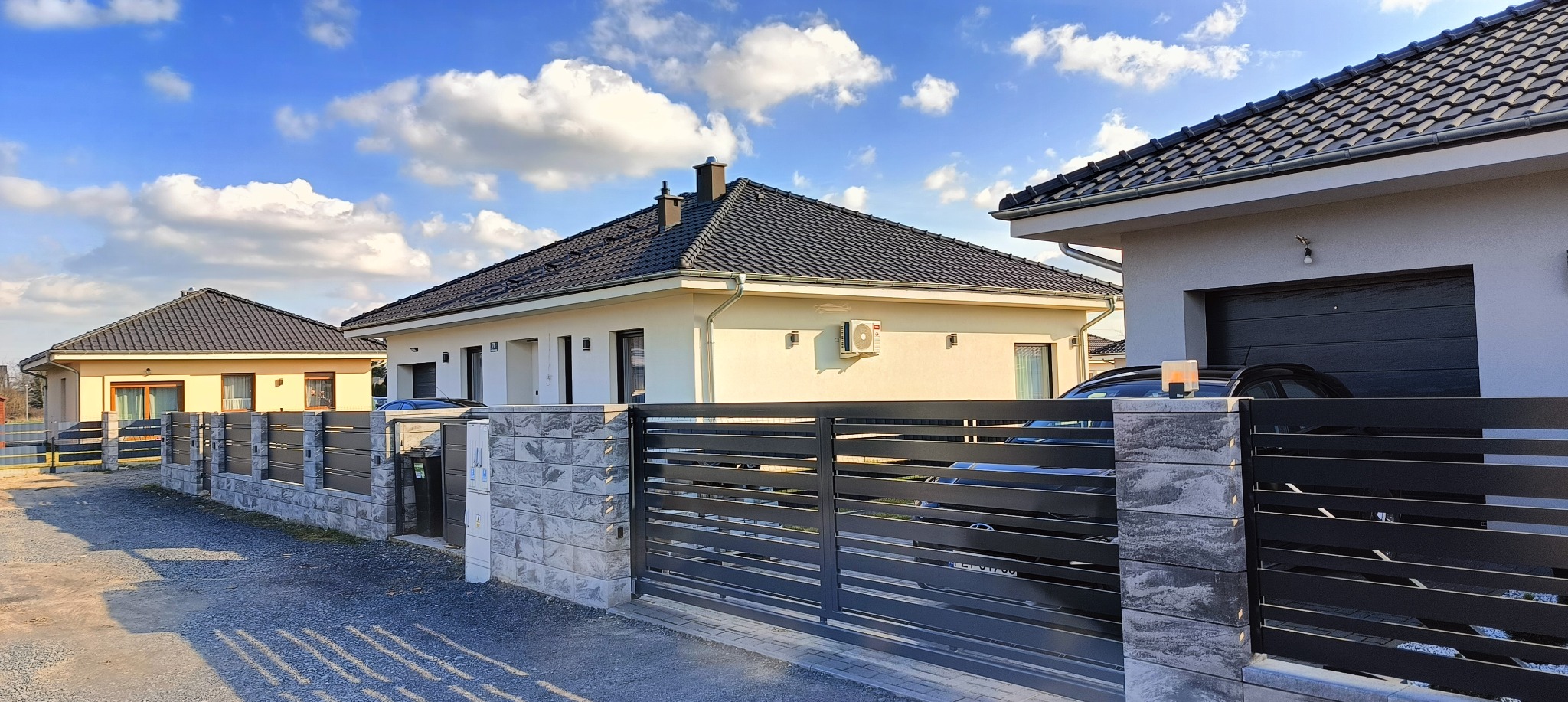 Nowoczesny dom jednorodzinny z klimatyzacją na elewacji, widoczny zza grafitowego ogrodzenia. Błękitne niebo z obłokami w tle. Słoneczny dzień.