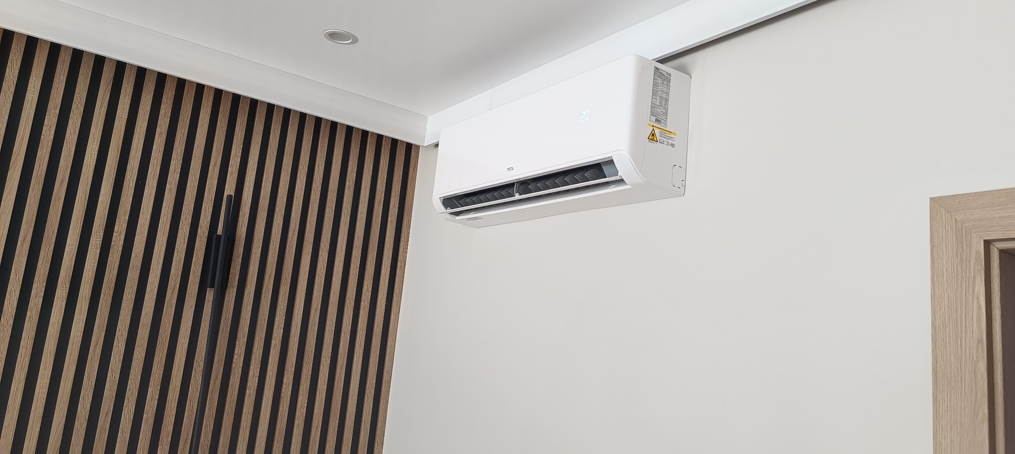 Biały klimatyzator TCL zamontowany na ścianie w nowoczesnym wnętrzu z dekoracyjną ścianą z drewnianych listew i fragmentem ramy drzwiowej.