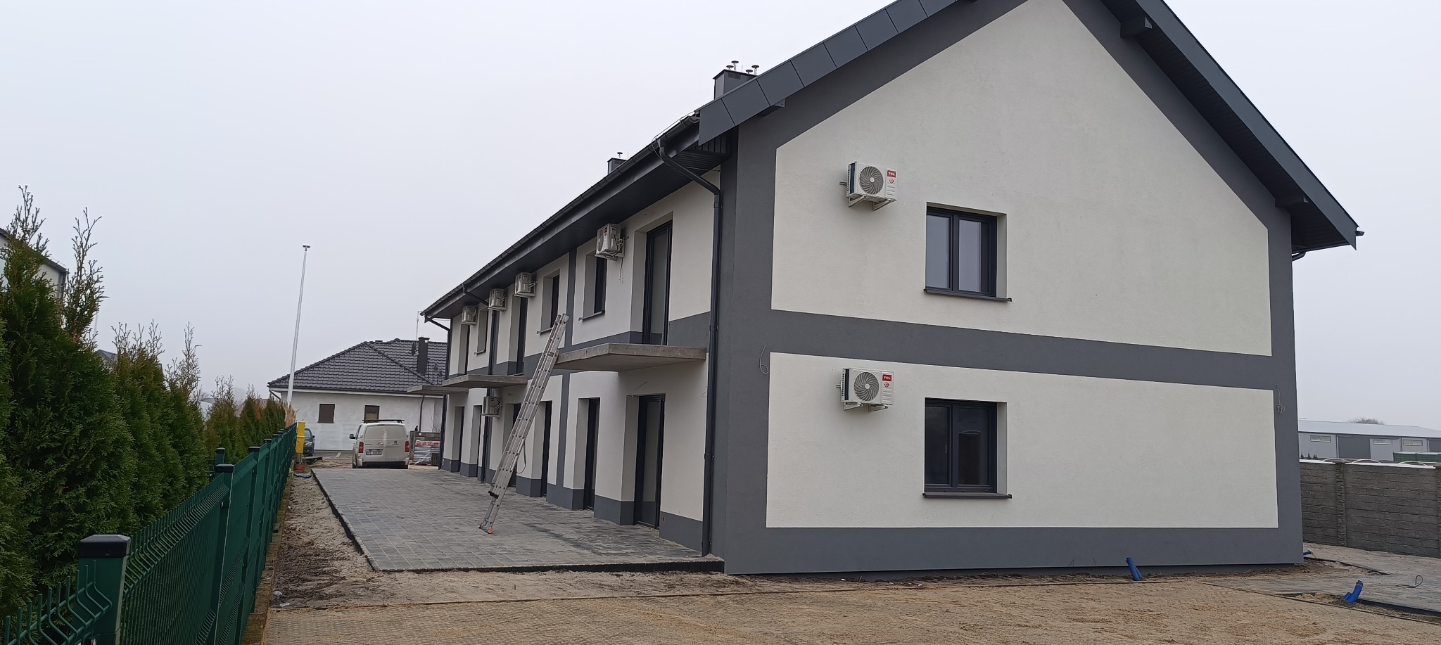 Szeregówka z zamontowanymi klimatyzatorami TCL na elewacji. Na pierwszym planie drabina oparta o budynek. Widok zewnętrzny, pochmurny dzień.