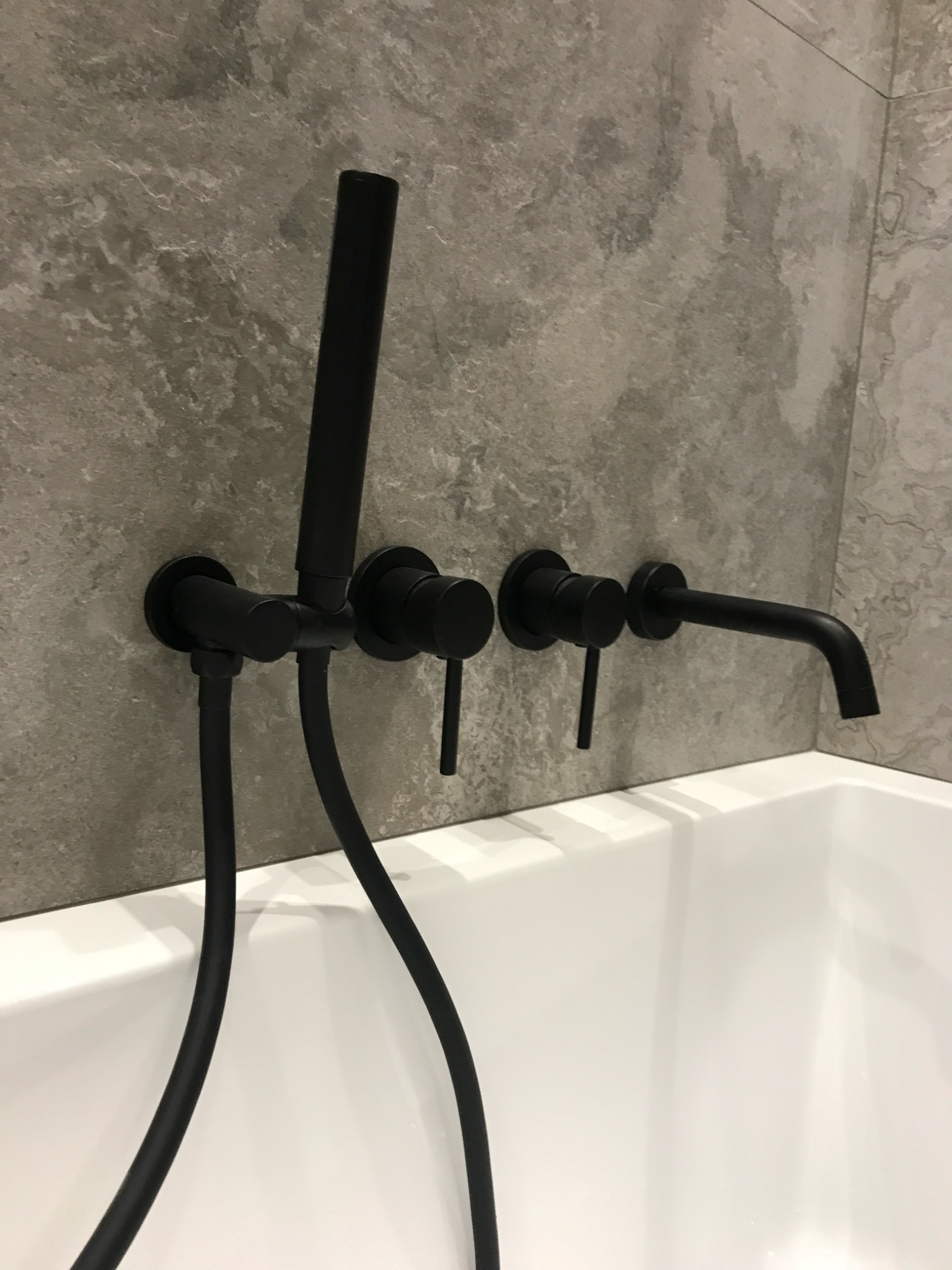 Minimalistyczna bateria wannowa w kolorze czarnym, zamontowana na szarej, marmurkowej ścianie. Nowoczesny design, widok z bliska na armaturę.