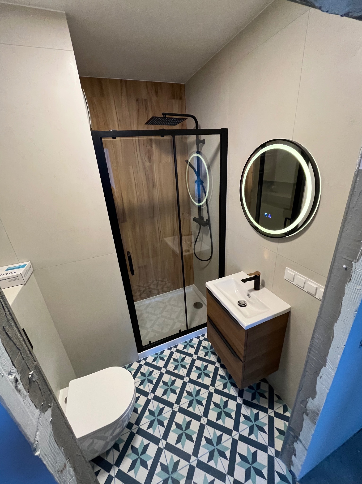 Nowoczesna łazienka po remoncie: kabina prysznicowa z czarną ramą, okrągłe lustro z podświetleniem LED, umywalka z szafką i wzorzyste płytki podłogowe.