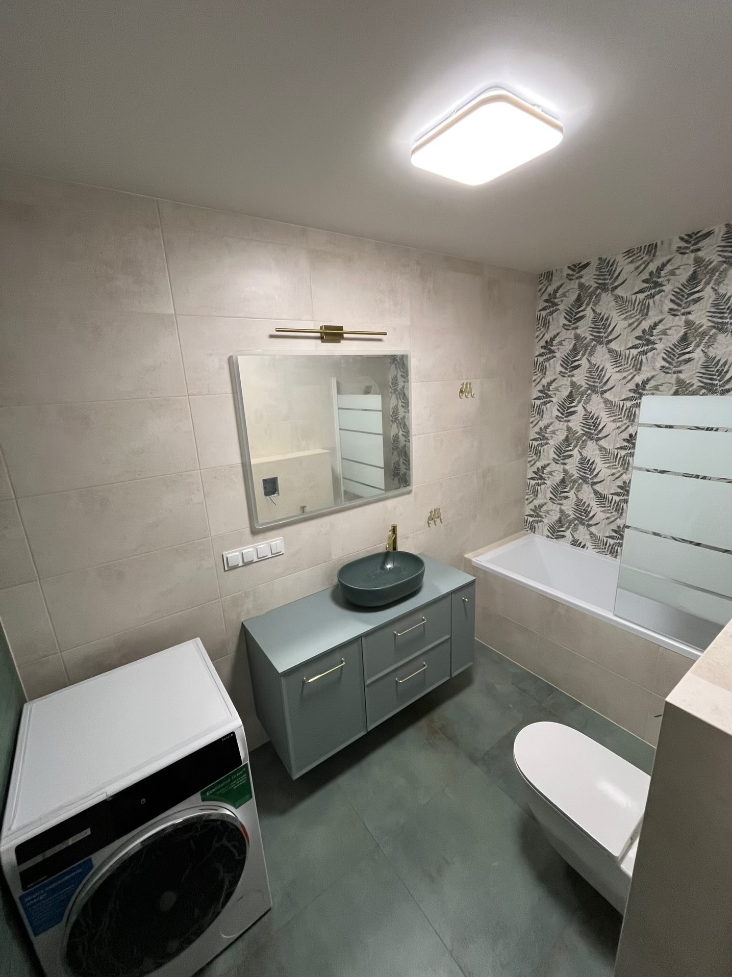 Wykończona łazienka z płytkami w odcieniach beżu i szarości, umywalką w kolorze butelkowej zieleni, pralką i toaletą. Na ścianie lustro z oświetleniem.