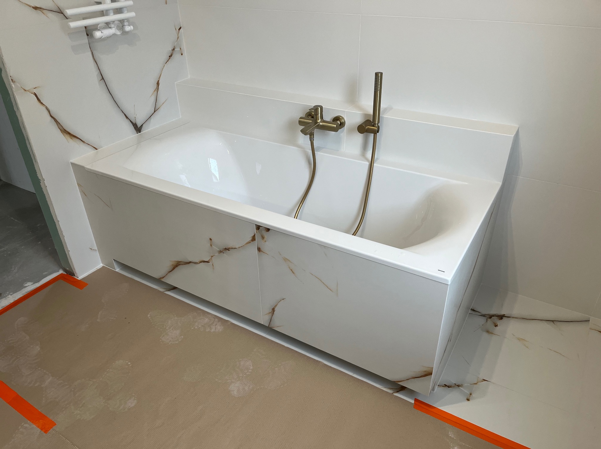 Biała wanna obudowana płytami imitującymi marmur z złotą armaturą. Widoczny fragment podłogi zabezpieczony kartonem i taśmą. Minimalistyczny design łazienki.