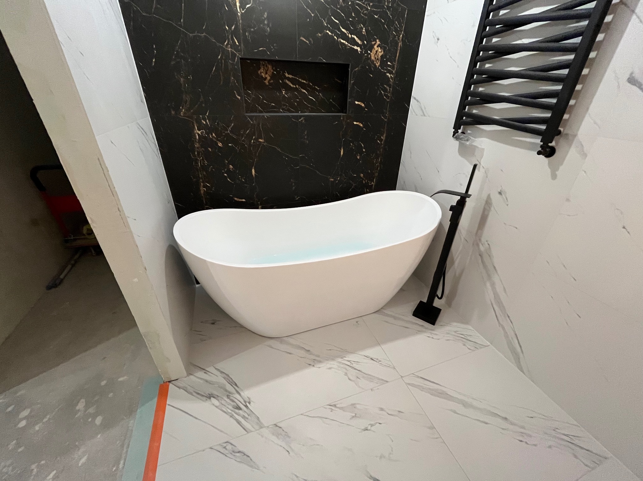 Nowoczesna łazienka z wolnostojącą wanną na tle marmurowych płytek i czarnej ściany z wnęką. Czarny grzejnik drabinkowy i bateria dopełniają elegancki wystrój.