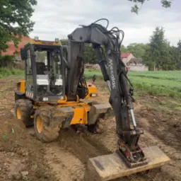Żółto-szara koparka Mecalac na błotnistym terenie, w tle zielone pole i domy. Widoczne ślady opon i świeżo przekopana ziemia. Pochmurne niebo.
