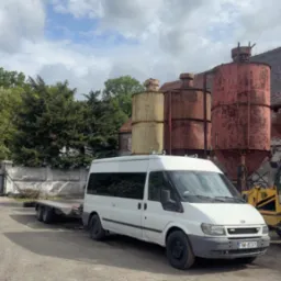 Biały bus z przyczepą lawetą na żwirowym placu, w tle silosy i budynek z cegły, częściowo widoczna koparka. Pochmurne niebo.