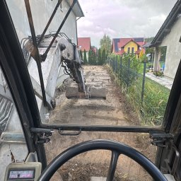 Obarewicz - Widok z kabiny koparki podczas prac ziemnych na działce, zarys fundamentów, domy w tle. Perspektywa operatora, pochmurne niebo.