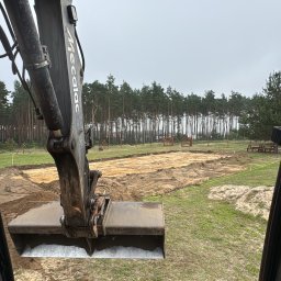 Obarewicz - Widok z kabiny koparki na przygotowany teren pod budowę, z widocznymi wykopami i lasem w tle, w pochmurny dzień. Ramię koparki z łyżką w kadrze.
