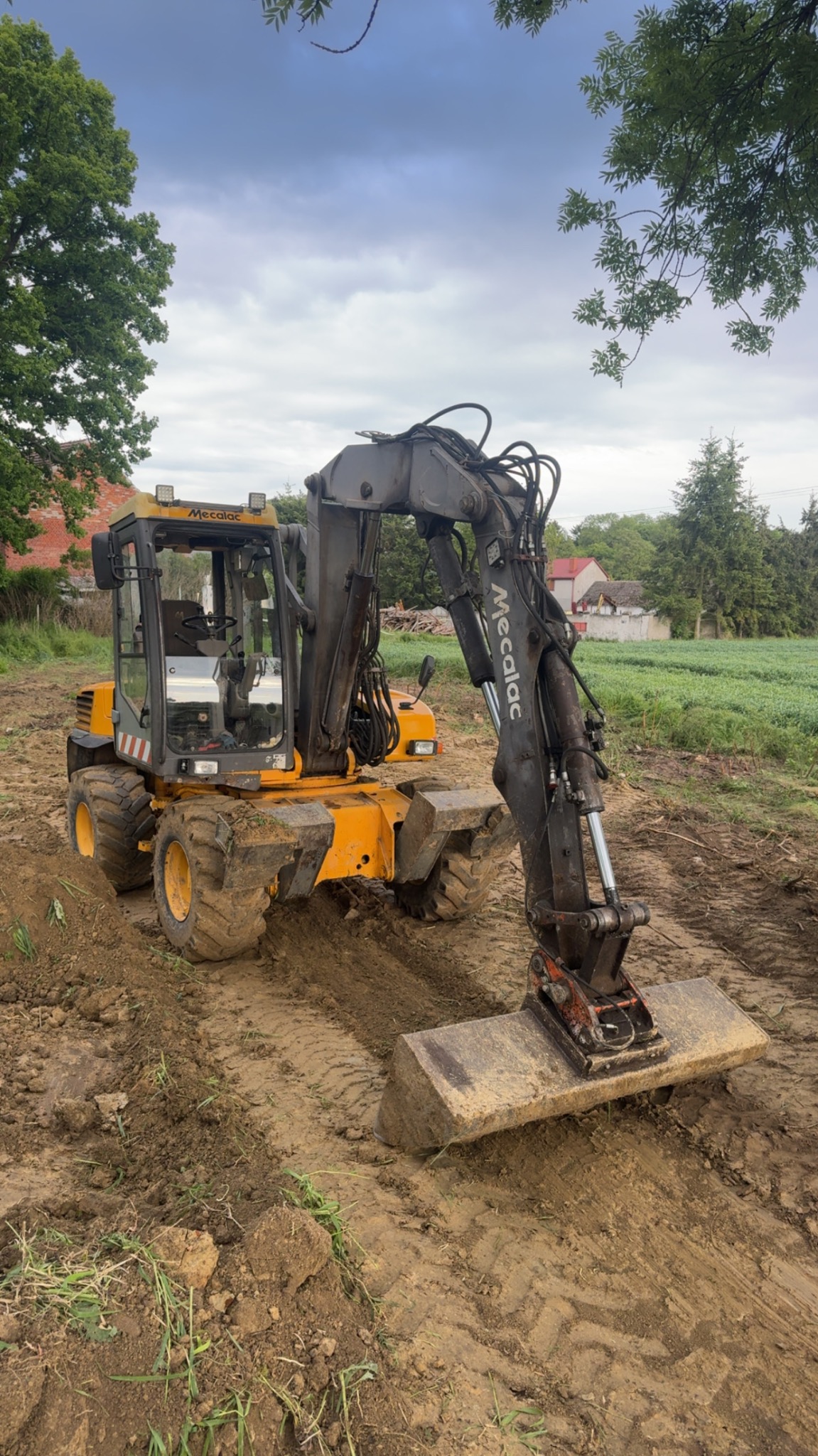 Żółto-szara koparka Mecalac na błotnistym terenie, w tle zielone pole i domy. Widoczne ślady opon i świeżo przekopana ziemia. Pochmurne niebo.