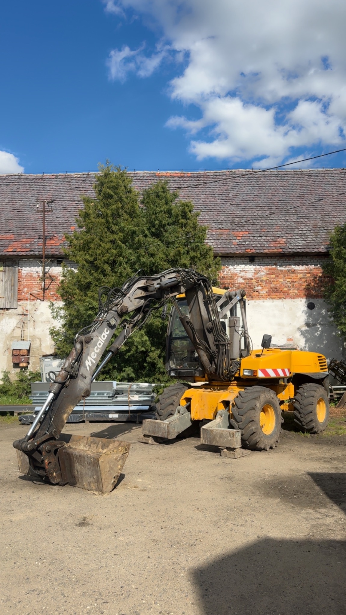 Żółta koparka Mecalac z łyżką na żwirowym placu, na tle budynku z cegły i drzew. Błękitne niebo z chmurami w tle. Maszyna gotowa do pracy.