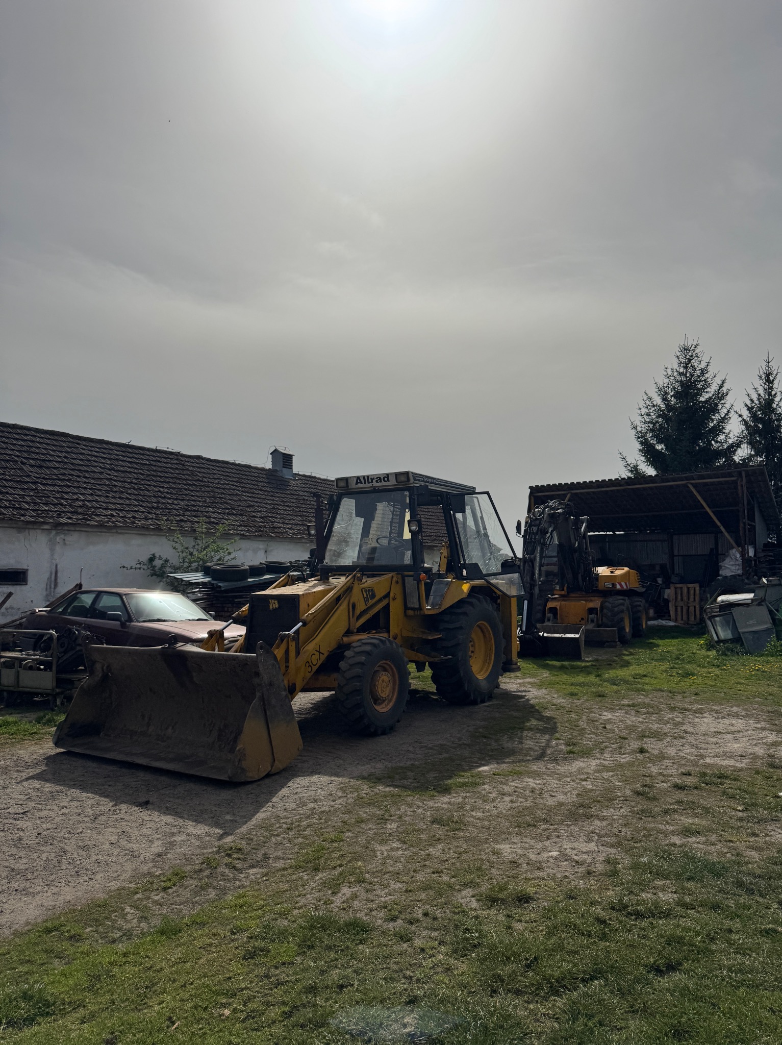Żółta koparko-ładowarka JCB 3CX na placu budowy, z widocznym łyżką i kabiną. W tle budynek gospodarczy i częściowo widoczna koparka.