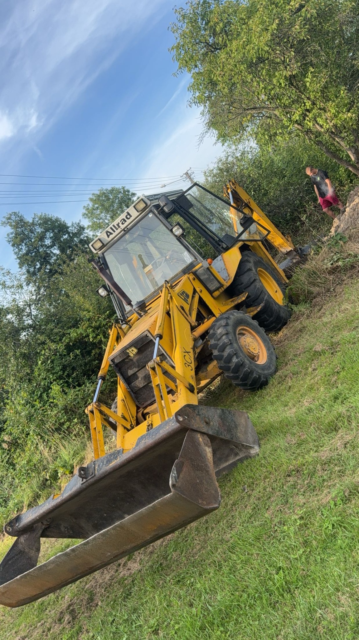 Żółta koparko-ładowarka JCB na trawiastym terenie, częściowo obrócona, z widoczną osobą w tle i błękitnym niebem. Maszyna gotowa do pracy.
