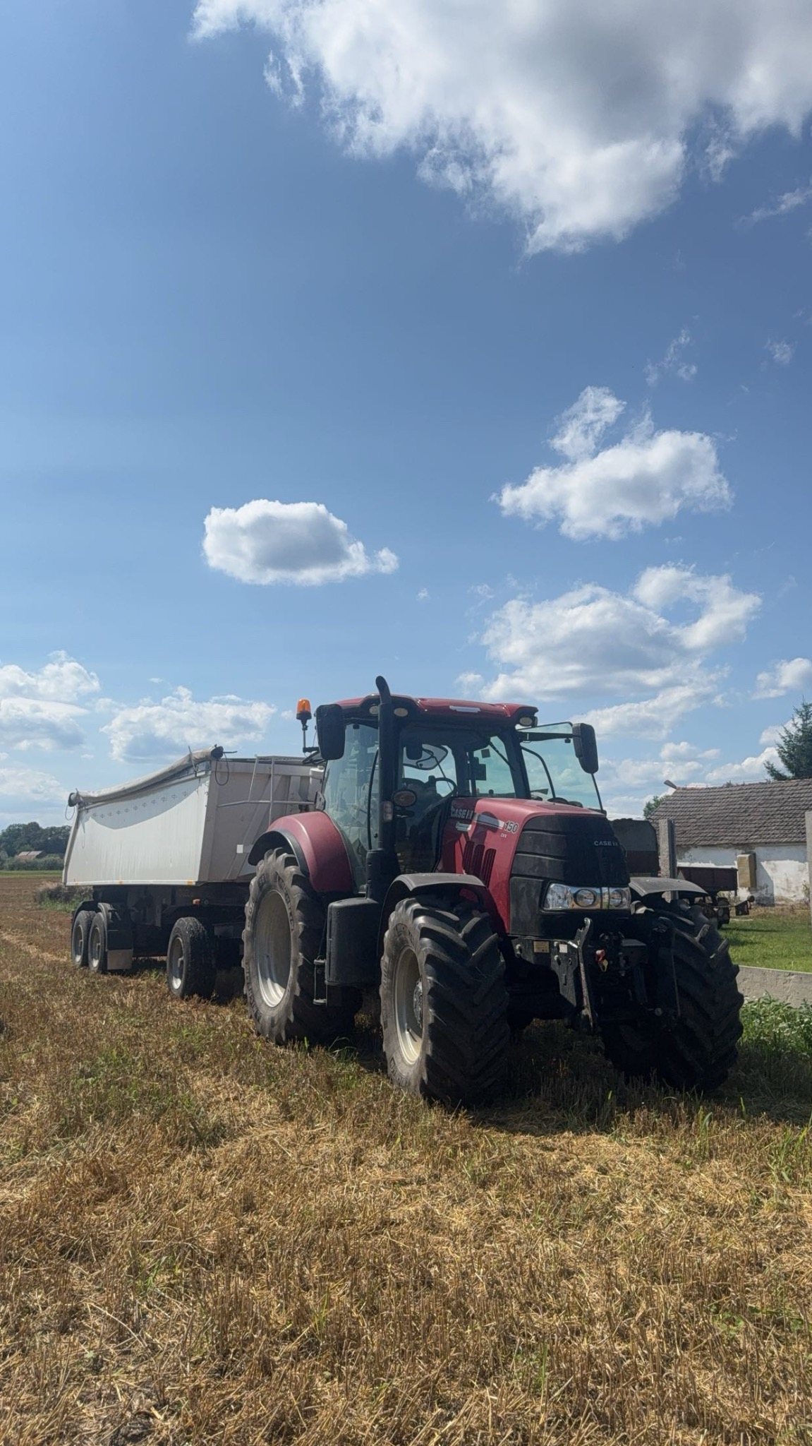 Czerwony traktor z przyczepą na polu w słoneczny dzień. Widoczne niebo z chmurami i budynek w tle. Transport materiałów rolniczych.